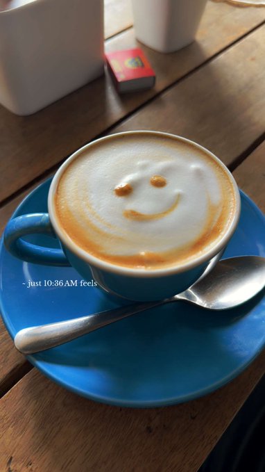 A blue coffee cup of latte with a smiley face drawn in the foam art, served on a matching blue saucer with a spoon, on a wooden table. Text overlay reads '- just 10:36 AM feels'
