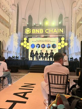 A conference stage with 'BNB CHAIN' branding and 'BNB' hexagonal displays where a panel of speakers is seated before an audience.