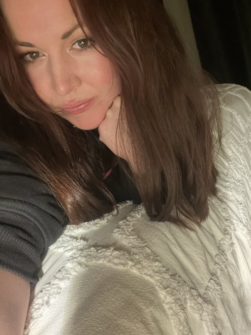 A woman with long brown hair rests her chin on her hand while wearing a black top, posed against a textured white blanket.