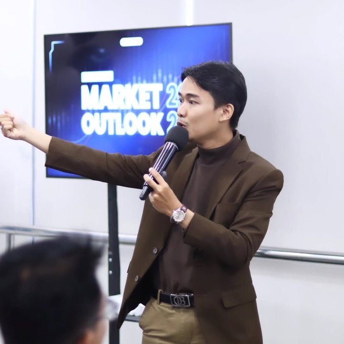 A man in a brown blazer speaks into a microphone at a "Market Outlook 2..." event, gesturing outward as he addresses an audience, embodying the post's humorous lament about speaking publicly yet remaining unrecognized without wealth or fame.