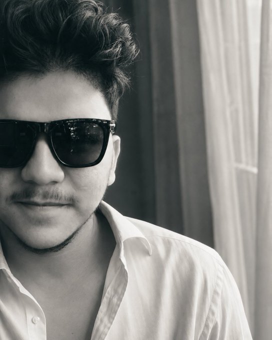 A black and white close-up portrait of a man with curly hair, a mustache, and sunglasses wearing a white button-up shirt.