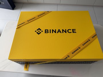 The image features a large, yellow box with a black "B" logo sitting on top of a white table. The box has a ribbon tied around its neck, adding an elegant touch to its appearance. The text "binance" can be seen in the image as well. The background is blurred and out of focus, drawing attention to the main subject - the yellow box with the black "B" logo on it.