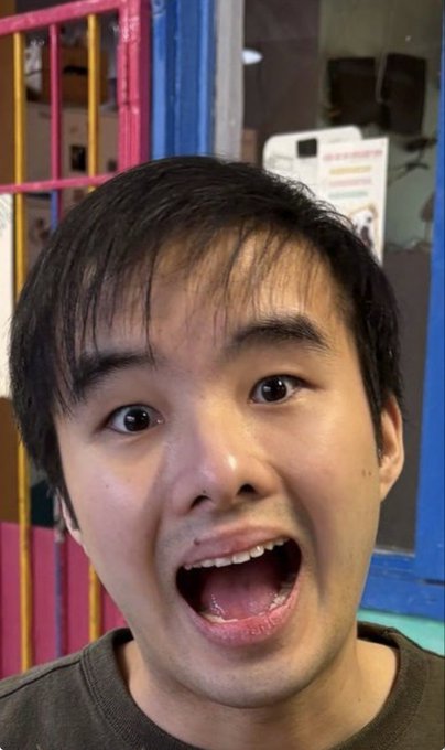 A close-up selfie of a young man with short black hair making a wide-open, exaggerated mouth expression, with colorful objects visible in the background.