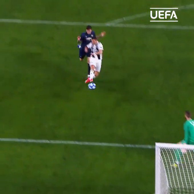 A still from a UEFA football match showing two players in mid-action on the green field, with one player in white challenging another in dark blue near the penalty area, while a goalkeeper in green stands ready by the goal.