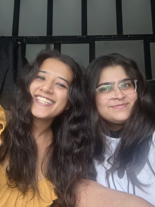 Two women pose closely together for a selfie—one with long dark wavy hair wearing a yellow top, and the other with glasses and a white t-shirt—smiling warmly in what appears to be a candid moment during the "Cowork with Maha" Bitget edition event in Bangalore.