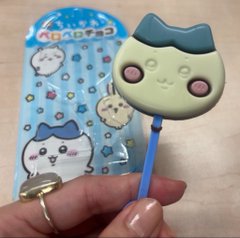 A person holds a small Chiikawa-themed item (a face-shaped clip or sticker) next to its original packaging featuring the cute character mascot.