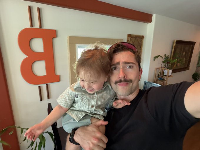 The image features a man holding a baby, both of them smiling for the camera. They are standing next to each other against a white wall with an orange sign that reads "Texas First $XMR Accepting Restaurant". The man appears to be wearing a black shirt and has a mustache.