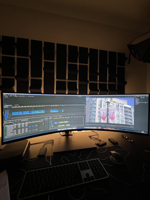 A dimly lit home office setup with dual curved monitors displaying video editing software on the left and a 3D modeling application on the right, with sound-dampening panels on the wall behind.