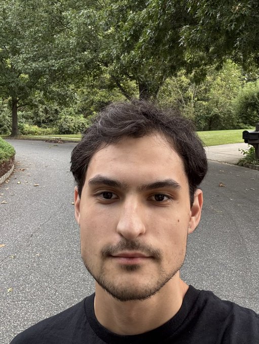 A close-up selfie of a young man with dark hair and a short beard wearing a black t-shirt, standing on a paved driveway with green trees in the background.