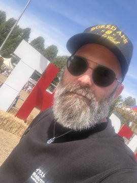 A bearded man wearing sunglasses and a black cap reading 'Naked Arms' takes a selfie outdoors near hay bales and a 'Pull' beer sign.