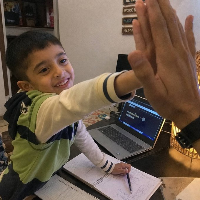 A photo of a young smiling boy at a desk giving a high-five to an adult's hand, with a laptop showing a Chrome browser and homework notebooks visible.