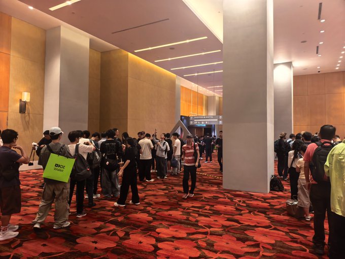 A crowded convention hall or hotel lobby with a red floral patterned carpet and many attendees wearing backpacks and event badges.