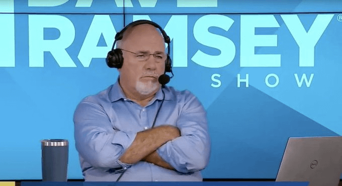 Dave Ramsey sits with arms crossed wearing a headset in front of 'THE DAVE RAMSEY SHOW' backdrop during a broadcast.