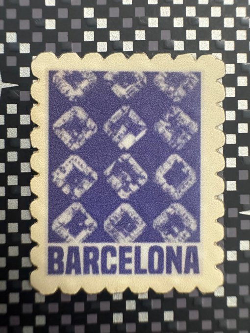 A vintage-style stamp or sticker with a purple checkered pattern and the word 'BARCELONA' printed at the bottom, placed on a black and silver patterned surface.