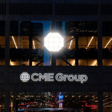 The image features a large building with a prominent CME Group sign displayed prominently above its entrance, indicating that the company operates within or around the city of New York. The building also has a large glass window reflecting the surrounding lights at night, creating an interesting visual effect. In addition to the main building, there is another smaller building visible in the background, suggesting that the area might be a commercial or financial district. A car can be seen driving by on the street below the building, and a person is standing near the entrance of the building, possibly a visitor or employee. The post also mentions the addition of altcoins like Avalanche and Cardano to the company's portfolio, indicating that CME Group is actively involved in the cryptocurrency market.