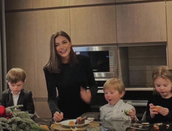 A woman in a black dress laughs while three young children eat cookies and snacks around a kitchen counter with a built-in microwave, with a festive holiday arrangement in the foreground. The humorous post jokes about the woman's "get rich quick" scheme involving a fictional "corgi" startup, playfully suggesting her wealth stems from posting dog selfies rather than any visible professional context in the cozy family scene.