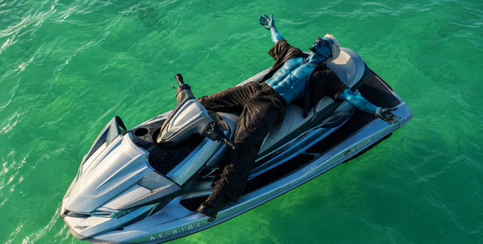 A blue-skinned humanoid figure in a black suit lounges across a jet ski floating on turquoise-green water.