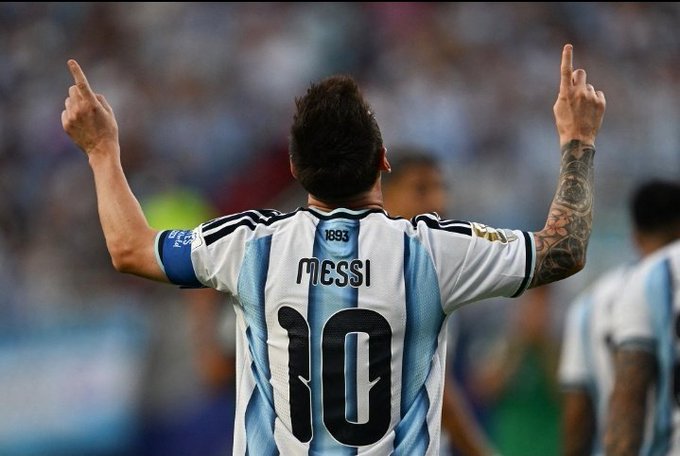 Lionel Messi, wearing the Argentina national team jersey with "MESSI" and the number 10, celebrates with both arms raised and index fingers pointed skyward, a gesture commonly associated with his goal celebrations. The image relates to the post as it showcases Messi after scoring one of his seven goals in the FIFA World Cup, the achievement being celebrated with the giveaway.