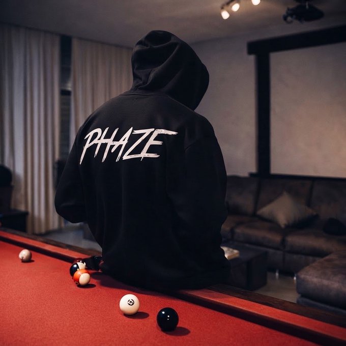 A person wearing a black 'PHAZE' hoodie sitting on the edge of a red pool table with several billiard balls scattered around, facing away toward a dark living room.