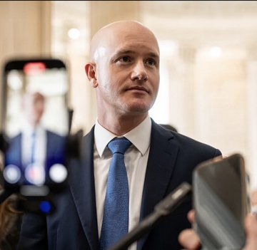 A bald man in a dark suit and blue tie speaks to reporters in what appears to be a government building hallway, with microphones and a smartphone recording him in the foreground. The image likely relates to the Coinbase executive advocating for the XRP Clarity Act, with the man likely being a Coinbase representative addressing the press about the national security concerns of delayed crypto legislation.