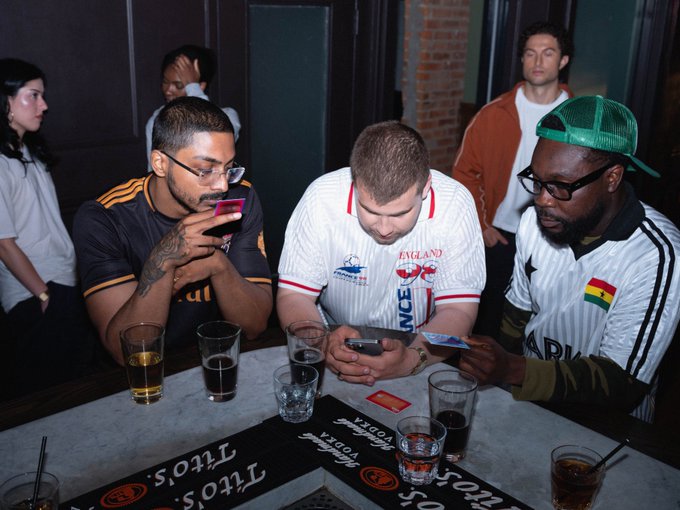 Three men sit at a bar looking at their phones, with the middle man wearing an England football jersey; the bar mat reads 'Linos'.