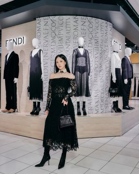 A woman with long dark hair poses in a black lace off-the-shoulder dress and black boots in front of a 'FENDI' retail display featuring mannequins dressed in elegant black and white attire.
