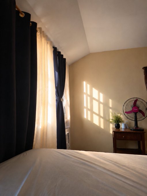 A sunlit bedroom corner features dark curtains framing a window, a wooden nightstand holding a vintage red fan and small plants, with sunlight casting a windowpane shadow on the beige wall.