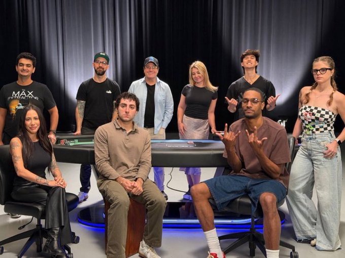 A group of eight casually dressed people pose around and behind a poker table in a studio with dark curtains, with one man seated on a wooden box making a hand gesture. A "MAX STAKES" logo is visible on the shirt of the man on the far left, and the poker table features "PokerStars" branding, suggesting this is a cast photo from the PokerStars creators content show—though the identities of the individuals remain unclear from the image alone.