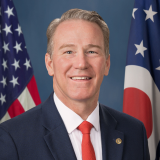 An official portrait of a man in a dark suit and red tie, standing in front of an American flag and another flag, likely a U.S. political figure.