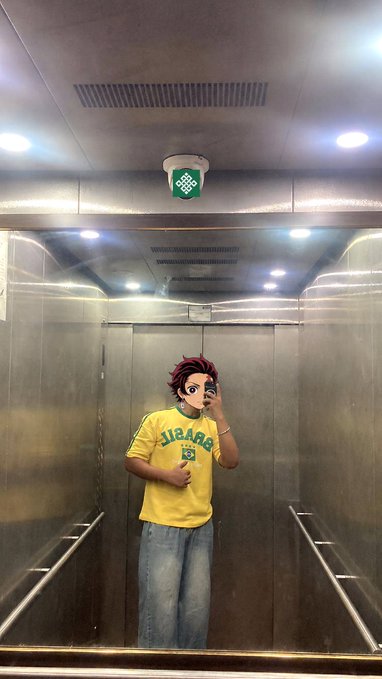 A person taking a mirror selfie inside a metallic elevator, wearing a yellow sweatshirt and with a digital anime-style mask covering their face.