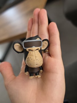 A small, handmade clay figurine of a Bored Ape-style NFT character wearing glasses and holding a beverage rests in the palm of someone's hand, showcasing a work-in-progress custom sculpture inspired by the user's NFT collection.