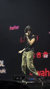 A concert scene showing a performer with dark hair on stage holding a microphone, with pink stage lighting and "!he!he" text visible on screens.