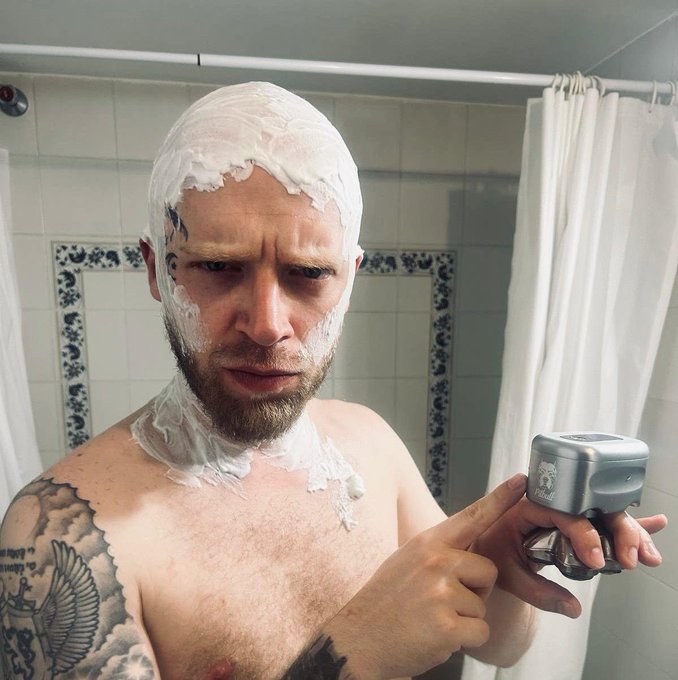 A shirtless man with a beard and tattoos, head covered in shaving cream, holds a Prilady razor while taking a mirror selfie in a bathroom. The image humorously illustrates the post's lament about losing his hair, capturing a candid "good morning" moment of grooming struggles.