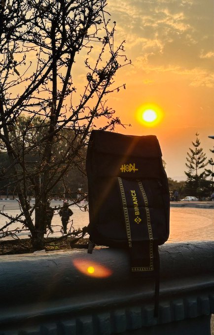 A black Binance-branded backpack rests on a railing silhouetted against a golden sunset, displaying "BINANCE" and the distinctive "ХОЯ" logo with a hidden "HOLD" message in the design. The image visually reinforces the post's theory that Binance has been subtly hinting at $MANYU through the recurring black-themed imagery and bag symbolism.