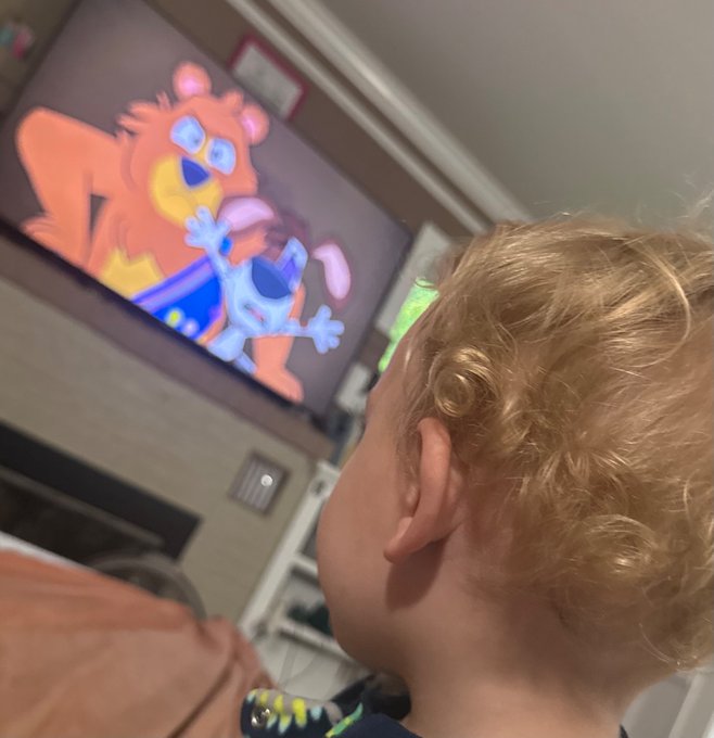 A young child with curly blonde hair is photographed from behind, looking up at a screen displaying a cartoon featuring an orange bear-like character and a blue creature.