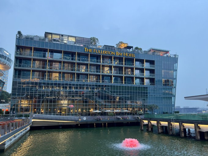 A dusk photograph of The Fullerton Bay Hotel, a modern glass-fronted building overlooking water with a pink illuminated fountain, with the Helix Bridge partially visible on the left.