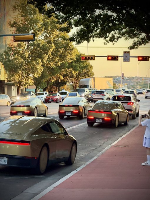 A line of sleek cars is stopped at a red traffic light on a city street, with a person standing on the sidewalk taking a photo. The scene has a nostalgic, film-like quality consistent with the post's claim that it is "Not an AI photo," featuring realistic traffic details and license plates that an actual camera would capture.