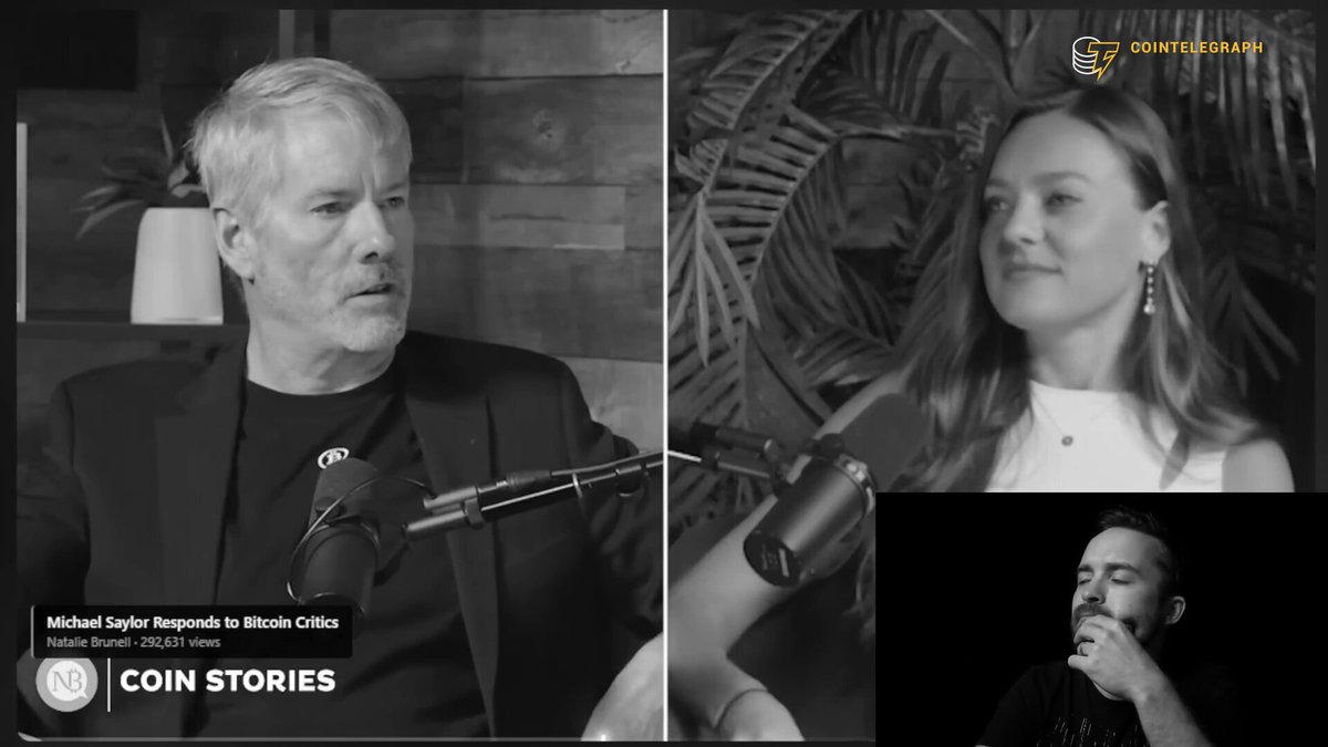 The image shows a man wearing a suit sitting behind a microphone, with another person seated across from him. They appear to be having a conversation or participating in an interview. The background features a wooden wall and a plant, creating a professional setting for their discussion. The text on the image reads "Michael Saylor responds to Bitcoin news at Coin Stories".