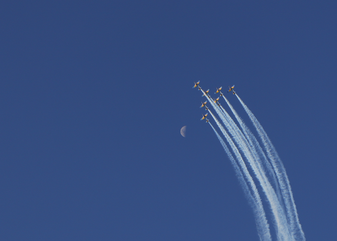 Five fighter jets fly in formation leaving white contrails against a deep blue sky, with a crescent moon visible in the background.