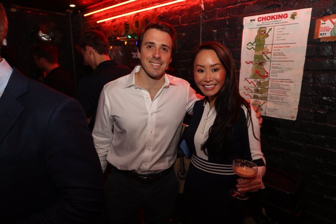 A man in a white shirt and a woman in a navy dress smile for a photo at a bar, with a 'CHOKING' first aid poster visible on the brick wall behind them.