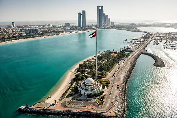 An aerial view of Abu Dhabi showing the iconic Etihad Towers skyline, a UAE flag, a domed monument, turquoise waters, and coastal roads.