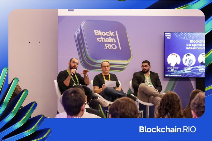 A panel discussion at the Blockchain.RIO conference featuring three men seated on stage with a purple 'Blockchain.RIO' logo backdrop and a screen displaying 'Blockchain: Dos agentes e Infraestrutura' with speakers Marcelo Creimer and Leonel Rampersad.