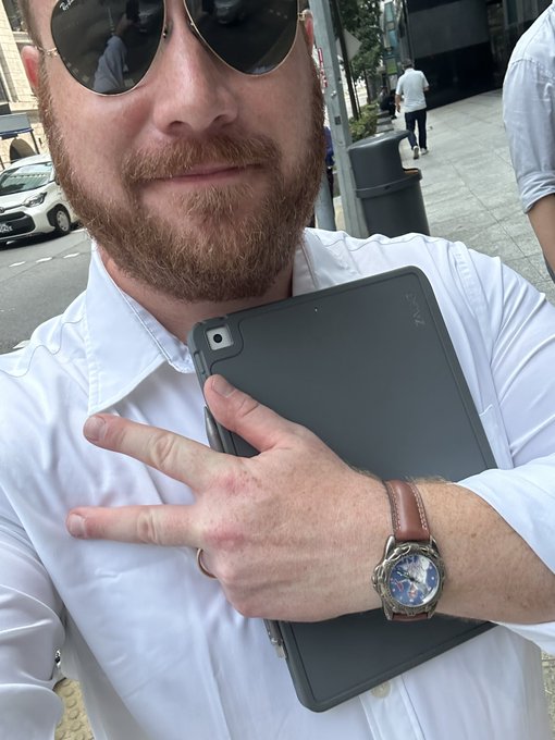 A selfie of a man with a reddish beard wearing aviator sunglasses and a white shirt, holding a tablet and wearing a watch.