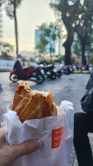 A hand holds a partially eaten sandwich (bánh mì) wrapped in paper with a 'THỔ NHĨ GIỐ' sticker, against a blurred street background with parked motorcycles and trees.