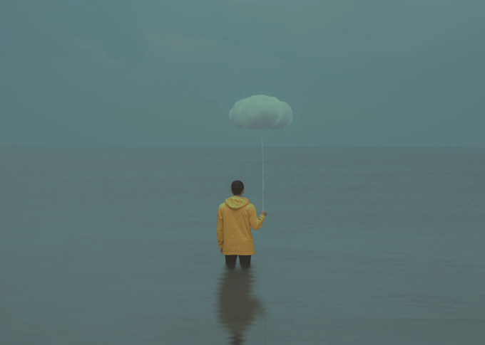A person in a yellow raincoat stands waist-deep in a vast, calm ocean holding a string attached to a small white cloud-shaped balloon against a hazy blue sky.