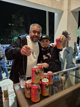 Two men pose together at what appears to be a nightlife event; one holds up a red can and several red cans are displayed on the table in front of them.