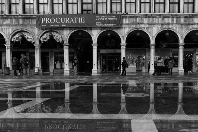 A black and white photograph of the Procuratie building featuring arched colonnades and a banner reading 'PROCURATIE - VISIT DISCOVER PLAY - THE HUMAN SAFETY NET A WORLD OF POTENTIAL', with people walking along a wet plaza reflecting the scene.