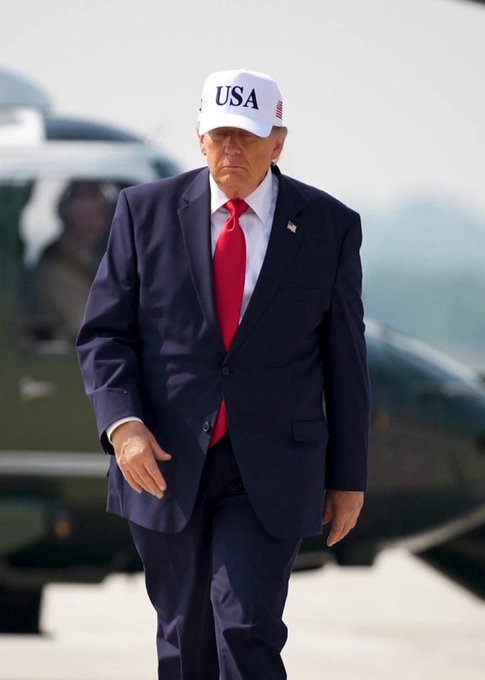 A man wearing a dark blue suit, red tie, and a white 'USA' cap walks in front of what appears to be a helicopter.