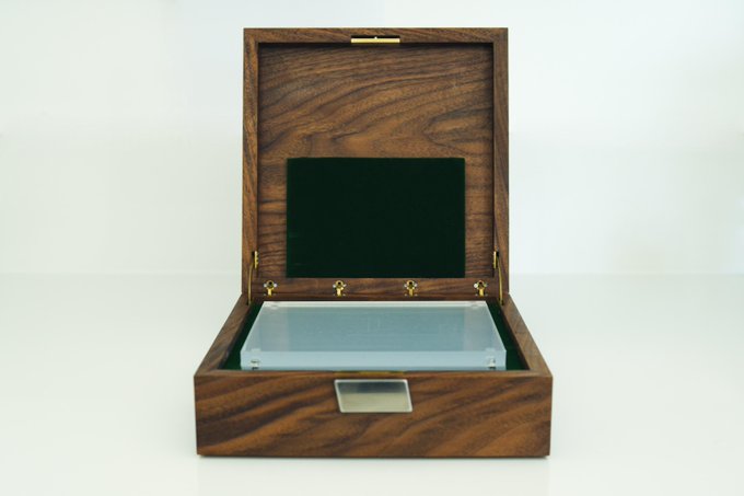 An open wooden display box with a green felt lining contains a clear acrylic case holding a silver coin or bar.