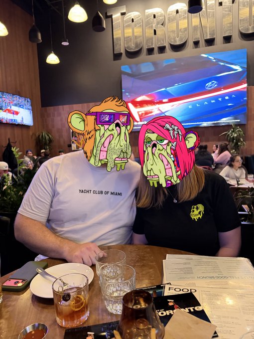 The image features a man and woman sitting at a table with drinks, including glasses of water and alcoholic beverages such as beer and wine. They are wearing funny masks that resemble cartoon characters or animals, adding a playful element to their gathering. The scene takes place in what appears to be an indoor setting, possibly a restaurant or bar.